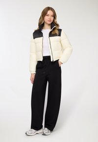 Giacca corta imbottita in crema e nero, con collo alto e chiusura con zip. Indossata con pantaloni neri a taglio ampio e sneakers argento.