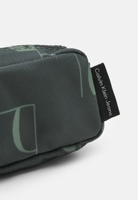 Pouch Calvin Klein Jeans in tessuto verde scuro con una superficie liscia, caratterizzato da discreti loghi e un'etichetta nera con branding.