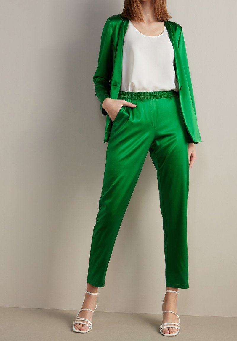 Costume en satin vert avec un blazer ajusté et un pantalon à taille élastique. Haut blanc sans manches. Talons blancs ouverts avec des lanières.