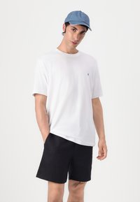 White cotton t-shirt with short sleeves and a small black logo, paired with black shorts. The person is wearing a light blue cap.