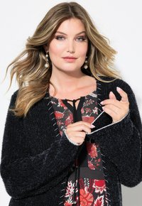 Mustapintainen pörröinen auki oleva neuletakki, jossa on valkoiset tikkaukset, käytetään kukkakuosisen mekon päällä. Malli pitää narua kädessään ja on pukeutunut helmikorvakoruihin.