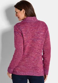 Vrouw gezien van achteren die een lange mouwen, roze-paarse gebreide trui en donkere broek draagt tegen een effen lichte achtergrond.