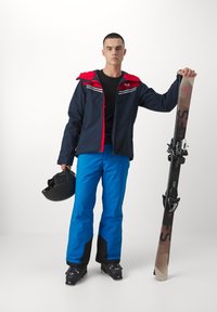 Skiausrüstung bestehend aus einer marineblauen Jacke mit roter Kapuze, blauen Hosen, einem schwarzen Helm und Skiern mit schwarzen Bindungen und einem gemusterten braunen Belag.