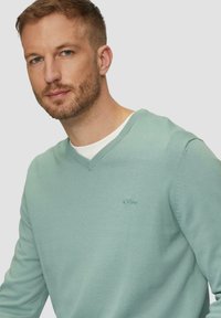 Heller grüner V-Ausschnitt-Pullover aus weichem Stoff, mit einem dezenten Logo auf der Brust, gerippten Bündchen und Saum, glatte Oberfläche.