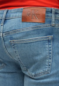 Šviesiai mėlyni džinsai iš džinso audinio, ant nugarinės juostelės su odine Calvin Klein logotipo etikete, standartinėmis galinėmis kišenėmis ir matomomis siūlėmis.