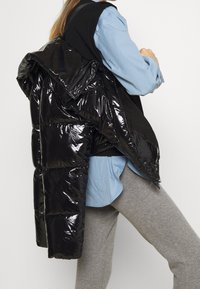 Sort, blank puffer jakke med hætte, lagd over en blå skjorte, parret med grå bukser. Jakken har quiltede sektioner og lynlås lukning.