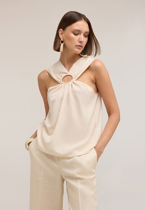 Frau mit glatten braunen Haaren trägt ein ärmelloses cremefarbenes Oberteil mit Schlüssellochausschnitt und passende cremefarbene Hosen, Hände in den Taschen, schaut nach unten.