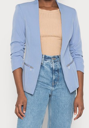 Kvinde iført en lyseblå blazer med rynkede ærmer, beige ribbet top og lyseblå jeans med høj talje, stående mod en ensfarvet baggrund.