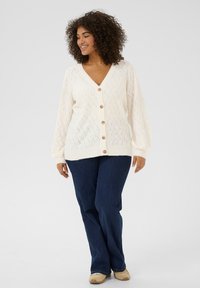 Cardigan crema a maglia con trama a cavo, scollo a V, chiusura anteriore con bottoni e maniche a palloncino, abbinato a jeans blu scuro e scarpe in tonalità neutra.