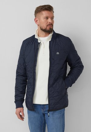 Mann trägt eine marineblaue gesteppte Jacke, einen weißen Pullover mit Reißverschluss bis zur Brust und blaue Jeans, steht mit einer Hand in der Tasche und schaut zur Seite.