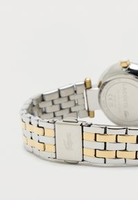 Hodinky ze stříbrného a zlatého kovu s náramkem na články. Hladký povrch s vyrytým logem na sponě. Kulatý ciferník s leštěným povrchem.