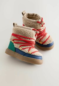 Kinderstiefel aus beigem,flauschigem Material mit roten Schnürsenkeln, blauen und grünen Akzenten, Gummisohle und einem gepolsterten Kragen für zusätzlichen Komfort.