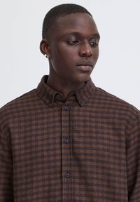 Jeune homme aux cheveux courts portant une chemise à carreaux marron et noire boutonnée, regardant vers le bas et vers sa droite sur un fond uni.