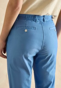 Niebieskie spodnie z elastycznym pasem o gładkiej fakturze; posiadają dwie tylne kieszenie z zapięciem na guziki. Zdjęcie wykonane z tyłu pod lekkim kątem.