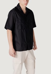 Camisa negra de manga corta con botones, dos bolsillos en el pecho, textura suave y cuello clásico, combinada con pantalones cargo de color crema.