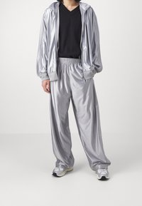 Osoba v lesklé stříbrné teplákové soupravě se zipovou bundou, černém tričku s výstřihem do V a bílými teniskami, stojící před jednobarevným světlem pozadím.