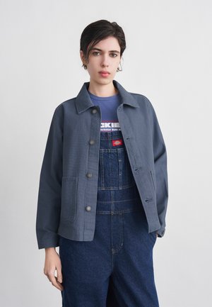 Młoda osoba o krótkich ciemnych włosach, ubrana w szarą kurtkę, niebieskie dżinsowe ogrodniczki i fioletowy T-shirt na jednolitym jasnym tle.