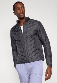 Black, padded puffer jacket featuring a high collar, zip closure, and side pockets. Textured design with contrast logo accent.