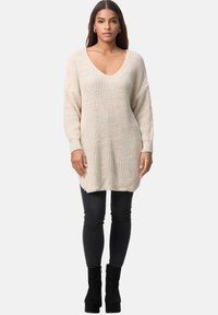 Beiger Strickpullover mit tiefem V-Ausschnitt, oversized Passform und gerippter Textur. Kombiniert mit dunklen Skinny-Jeans und Ankle Boots.