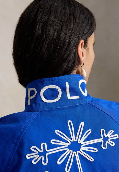 Persoon met donker haar draagt een blauw shirt met 'POLO' geborduurd op de kraag en een wit abstract florale ontwerp op de achterkant.