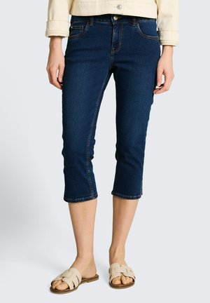 Kobieta w ciemnoniebieskich krótkich dżinsach, beżowej kurtce i beżowych sandałach wsuwanych, stojąca na jasnym, jednolitym tle.