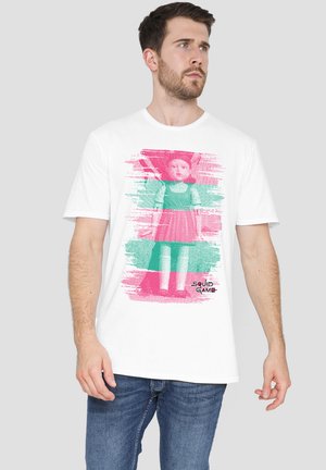White cotton t-shirt featuring a pixelated graphic of a doll in a pink dress, with green and pink brushstroke accents. Text below reads "SQUID GAME."
