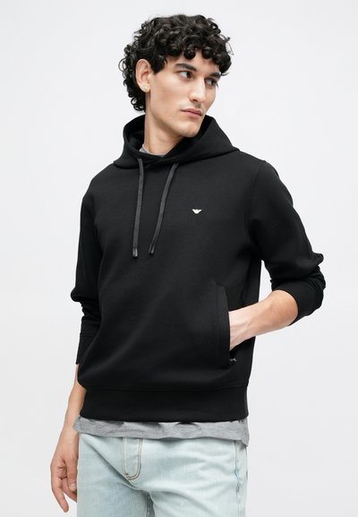 Melns džemperis ar kapuci, priekšējā kabata un auklu detaļa, izgatavots no gludas auduma. Uz krūtīm ir mazs logotipa akcenta elements un riboti aproces gali.