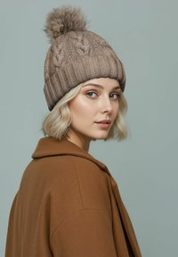 Knit beige beanie with a textured pattern and a fur pom-pom on top, worn with a brown coat against a light green background.