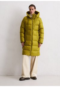 Lange pufferjas in olijfgroen, met een ritsluiting, capuchon en een boxy gewatteerd ontwerp. Gepaard met brede crèmekleurige broek en bruine chunky schoenen.