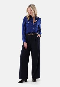Blue satin shirt with two chest pockets, button closure, and long sleeves. Paired with high-waisted black wide-leg trousers and heeled boots.