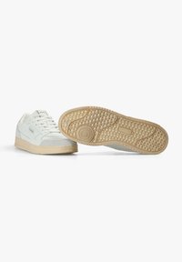 Zapatillas blancas hechas de lona y ante, con una suela de goma beige que presenta un patrón texturizado y un detalle de logo redondo en la suela.