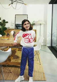 Un pull blanc avec un motif de cerises rouges, un pantalon de sport bleu avec des rayures blanches et des baskets blanches. Cadre intérieur avec une table en bois et des plantes.