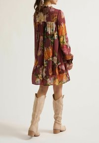 Model in een bloemenjurk met lange mouwen en geplooide zoom, met kniehoge beige laarzen, van achteren gekeerd tegen een effen achtergrond.