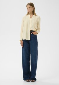 Crème knoopshirt met verticale plooien, gecombineerd met hooggezette donkerblauwe wijde jeans. Model staat met de handen in de zakken.