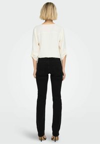 Top blanco de manga larga con puños enrollados, combinado con jeans negros ajustados y tacones negros, visto desde atrás sobre un fondo claro.