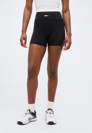 Personne athlétique portant un short noir taille haute Reebok, un haut court blanc, des chaussettes blanches et des baskets blanches Reebok sur un fond uni.