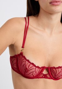 Red lace bra with sheer mesh cups, scalloped edges, and double straps. Notable gold hardware detail at the center.