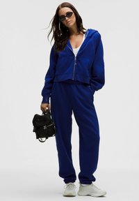 Felpa corta blu con tasche frontali, abbinata a pantaloni jogger coordinati. L'outfit ha una texture morbida ed è completato da sneakers bianche e uno zaino nero.