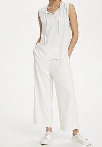 Blusa bianca senza maniche con dettagli in pizzo sul colletto, abbinata a pantaloni bianchi larghi e sneakers bianche.