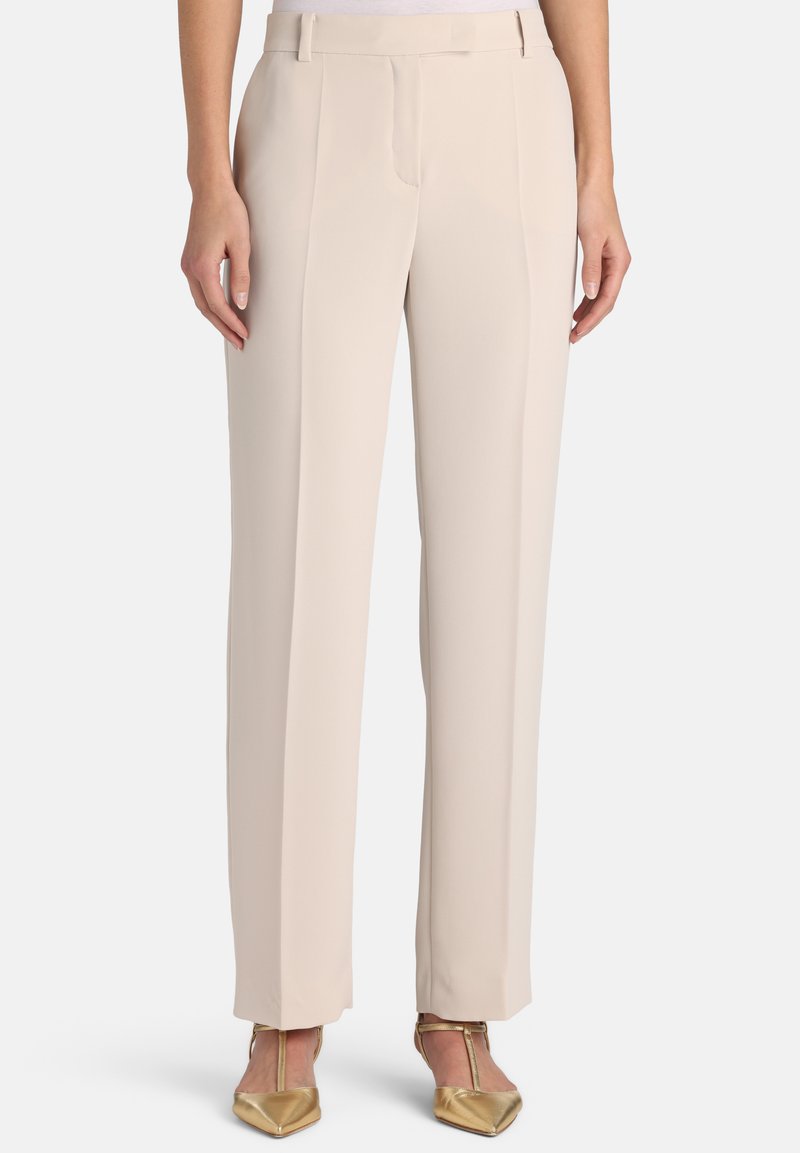 Pantalon beige sur mesure avec devant plat, jambes repassées et tissu lisse et léger ; porté avec des chaussures dorées à bout pointu.
