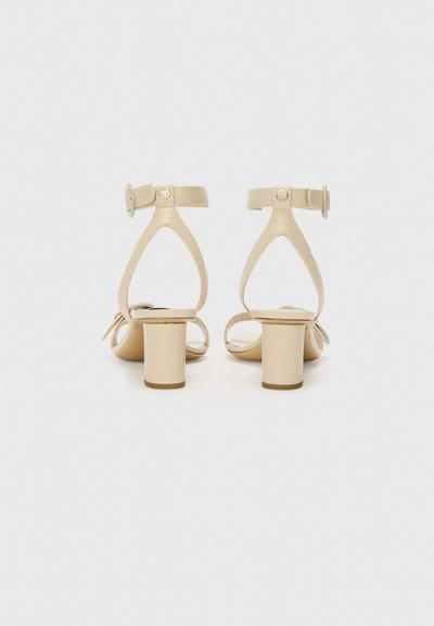 Beige højhælede sandaler med en kraftig hæl, åben bag, og ankelrem med en cirkulær spænde. Glat læderoverflade, minimalistisk design.