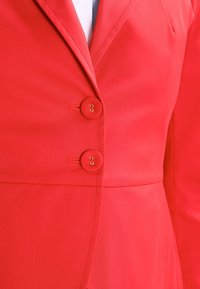 Blazer rouge avec boutons dorés, col structuré, tissu lisse et coupe ajustée avec des détails subtils de couture.