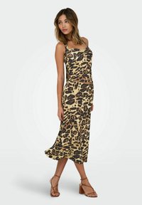 Leopard print midi-kjole i beige og sort, med tynde stropper og en tætsiddende overdel, der flyder ud i en løs nederdel. Lysebrune strop sandaler.