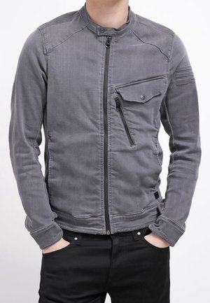 Homme portant une veste grise ajustée zippée avec une poche poitrine inclinée et un pantalon noir, mains dans les poches, recadré sous le menton.