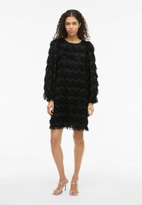 Robe noire avec un motif en vagues texturé et des détails en franges, à manches longues et coupe droite, accompagnée de sandales à talons argentées.