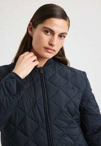 Giovane donna con lunghi capelli scuri indossa una giacca nera trapuntata con zip, toccando delicatamente il colletto su uno sfondo chiaro e semplice.