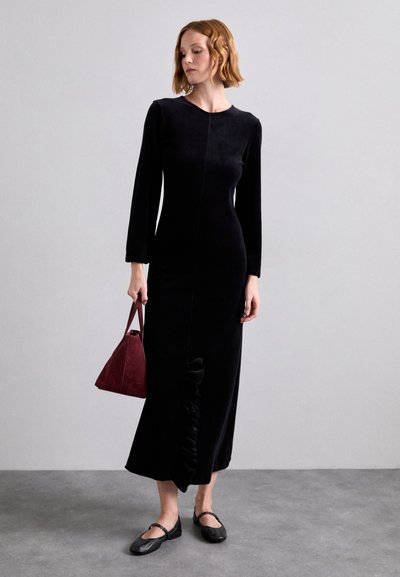 Black velvet long-sleeve dress with a fitted silhouette, side ruffle detail, paired with a maroon triangular handbag and black flats.
