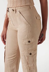 Beige Cargo-Hosen aus leichtem Material mit mehreren Taschen, Reißverschlussdetails und strukturierter Oberfläche. Verfügt über einen verstellbaren Bund.