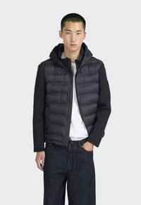 Giacca a vento nera con cappuccio e maniche in pile, con chiusura a zip, texture trapuntata e vestibilità casual, indossata sopra una maglietta bianca.