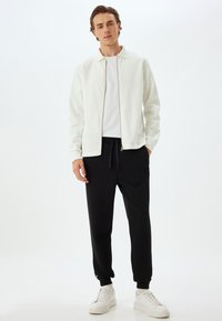 Chaqueta blanca con cremallera sobre una camiseta blanca, combinada con pantalones de chándal negros y zapatillas blancas. Tejido suave, corte relajado, detalles sutiles.